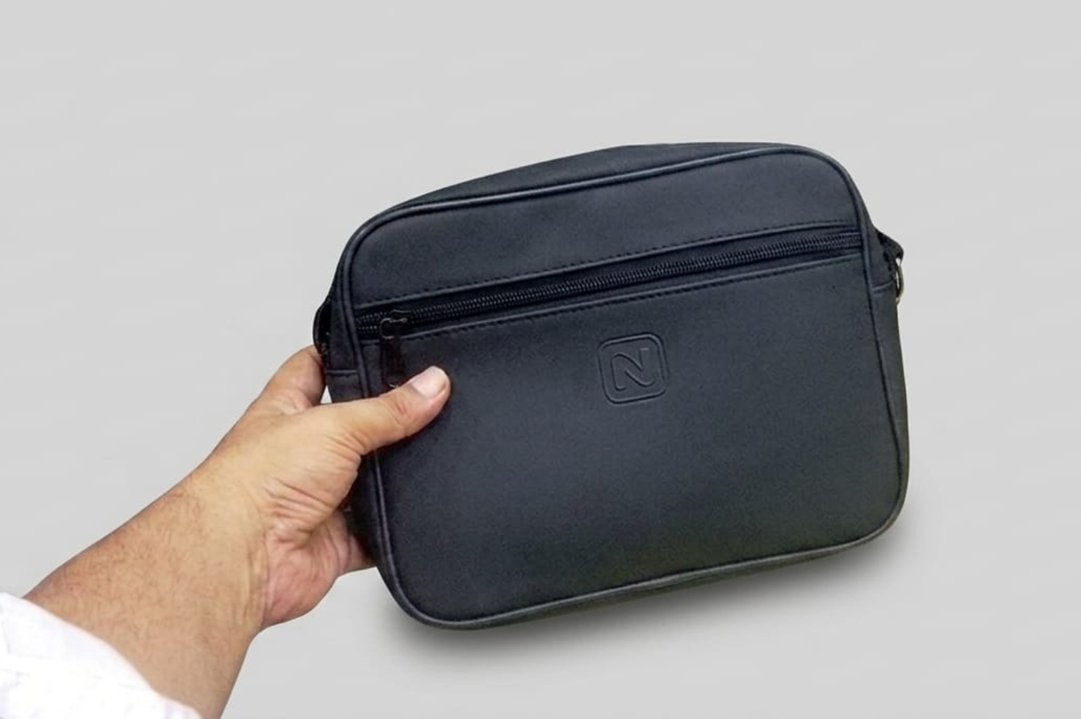 Hand holding a black leather briefcase with a logo on a gray background